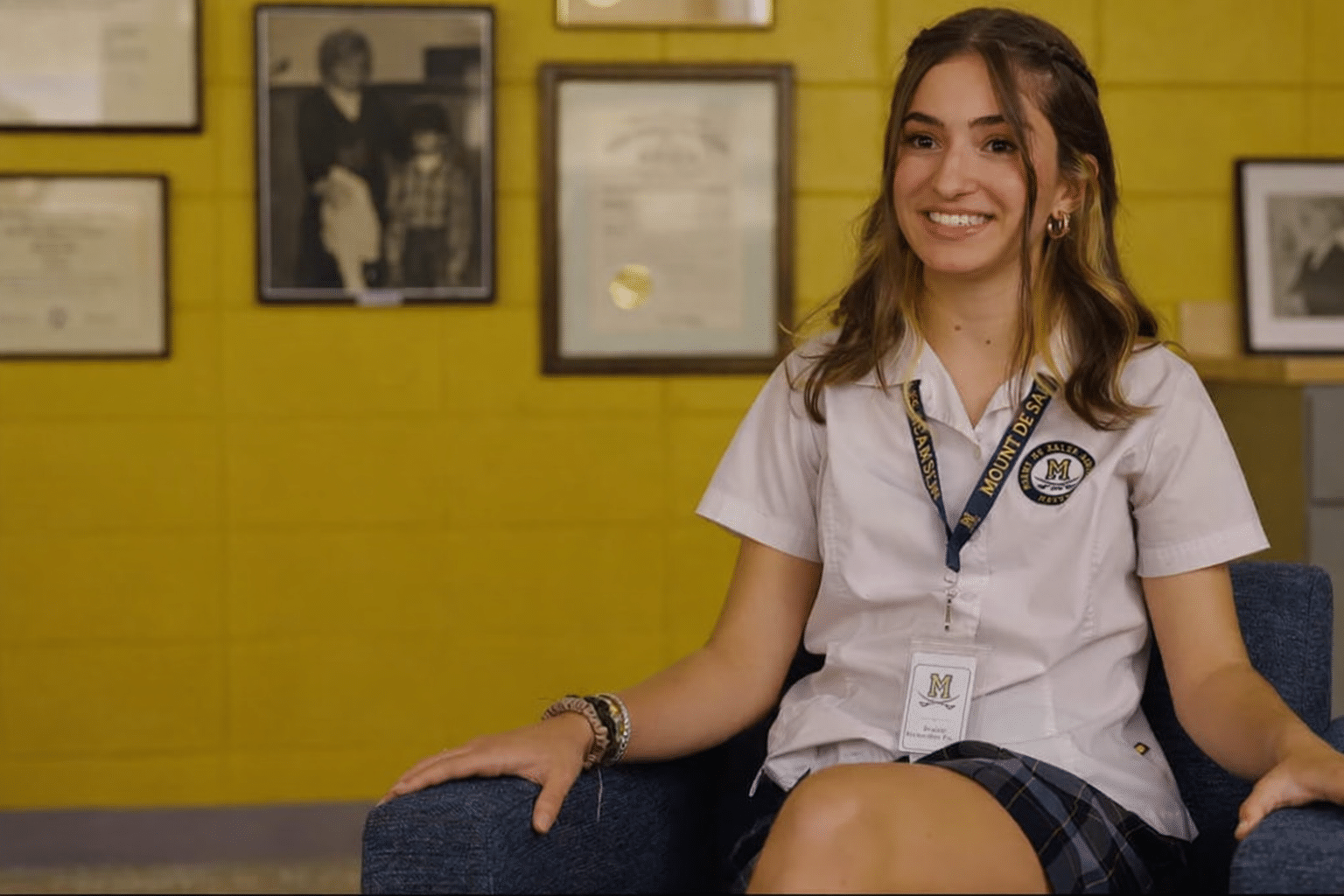 A cheerful teenage girl in a school uniform and lanyard sits on a chair in front of a yellow wall with framed photos and certificates.