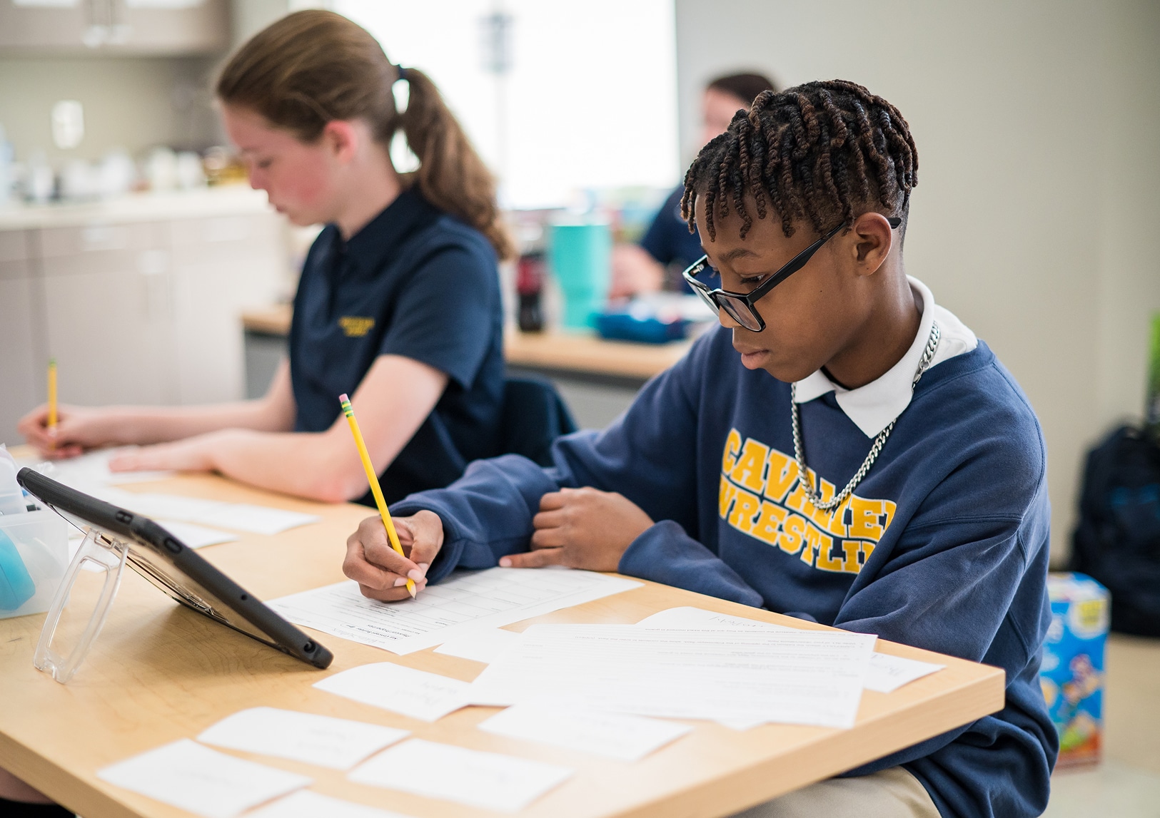 Two students sit at a desk working on assignments. One writes on paper with note cards, glasses, Cavaliers Wrestling sweatshirt. The other writes and uses a tablet.