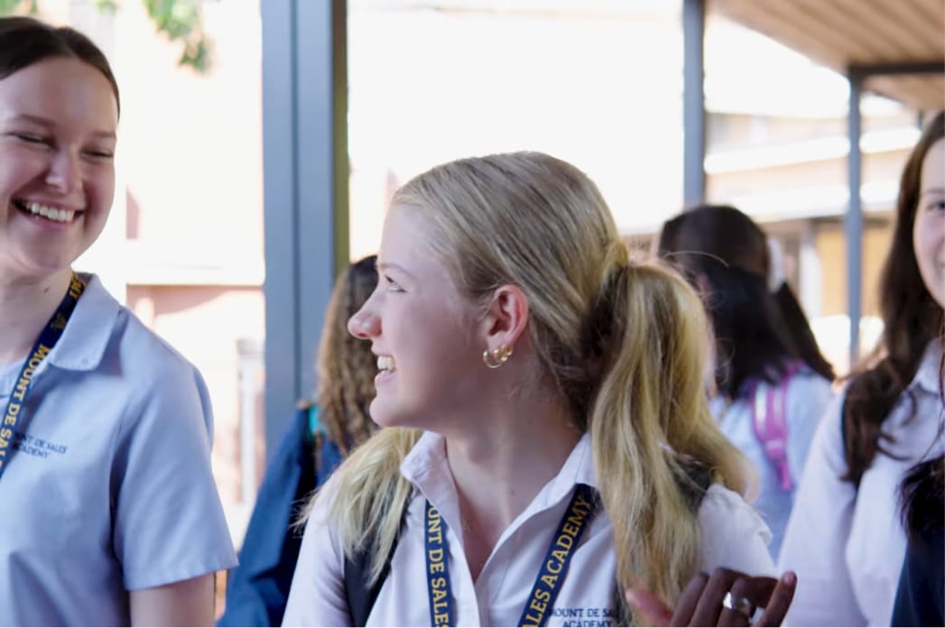 Three students in school uniforms smile and chat outdoors, wearing lanyards. More students and a covered walkway are visible behind them.